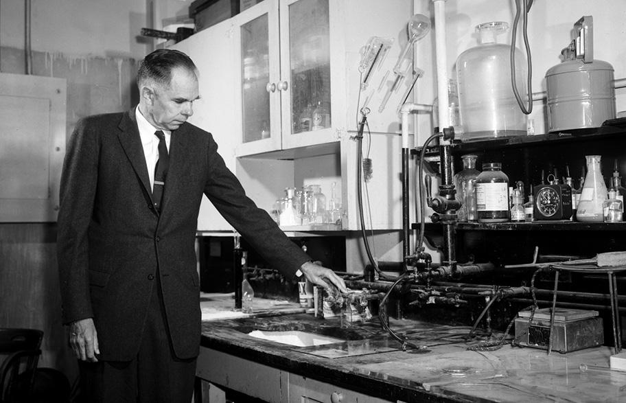 Mustavalkoinen valokuva, jossa miesoletettu puvussa seisoo laboratorion pöydän ääressä ja katsoo jonkinlaista laitetta.