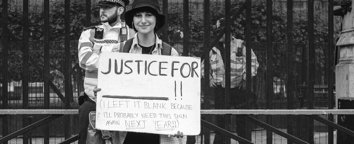Henkilö seisoo kadulla korkean aidan edessä ja pitää käsissään kylttiä, jossa lukee "Justice for - ! (I left blank because I'll probably need this sign again next year!)". Henkilön takana seisoo poliisi ympäristöä tarkkailemassa.